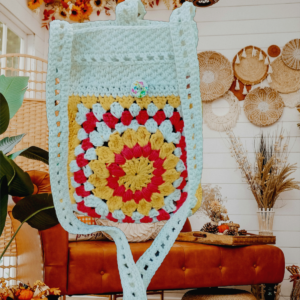 “Pochette granny square crochetée main en coton recyclé blanc cassé, rouge et jaune tournesol, avec bandoulière de 120 cm et fermeture par bouton en bois naturel.”