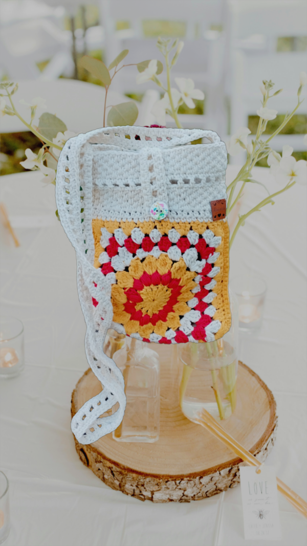 “Pochette granny square crochetée main en coton recyclé blanc cassé, rouge et jaune tournesol, avec bandoulière de 120 cm et fermeture par bouton en bois naturel.”