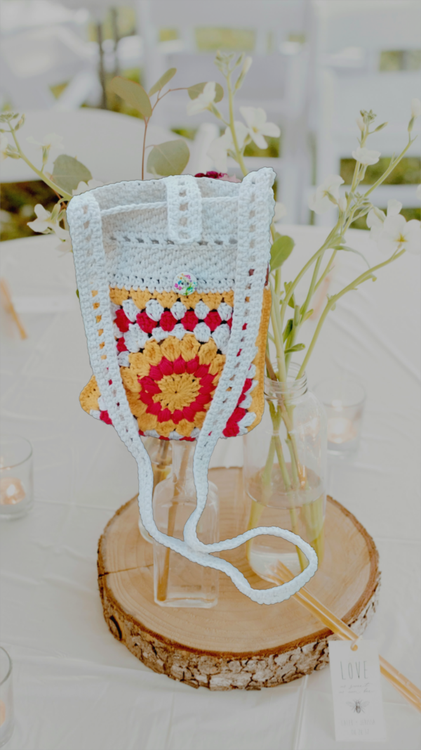 “Pochette granny square crochetée main en coton recyclé blanc cassé, rouge et jaune tournesol, avec bandoulière de 120 cm et fermeture par bouton en bois naturel.”