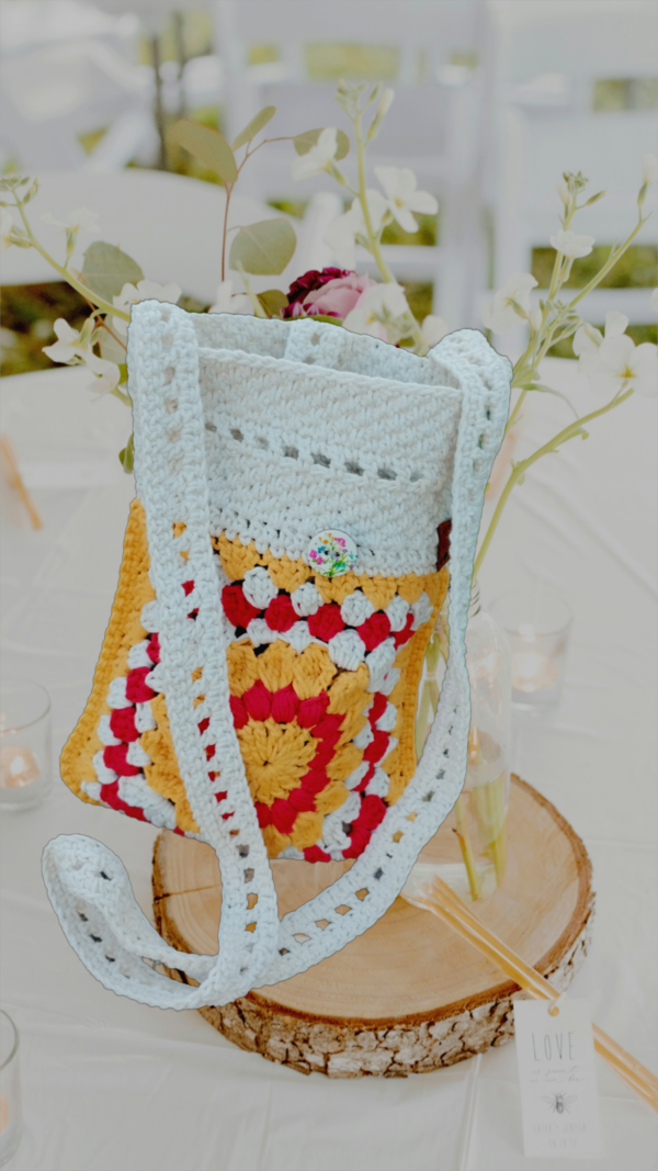 “Pochette granny square crochetée main en coton recyclé blanc cassé, rouge et jaune tournesol, avec bandoulière de 120 cm et fermeture par bouton en bois naturel.”