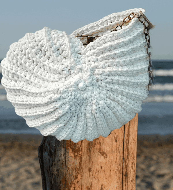 Sac crocheté à la main en coton recyclé, forme inspirée des coquillages, avec une chaîne en métal et une perle nacrée en ornement.