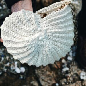 Sac crocheté à la main en coton recyclé, forme inspirée des coquillages, avec une chaîne en métal et une perle nacrée en ornement.