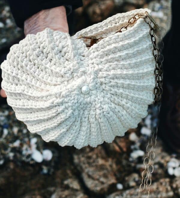 Sac crocheté à la main en coton recyclé, forme inspirée des coquillages, avec une chaîne en métal et une perle nacrée en ornement.