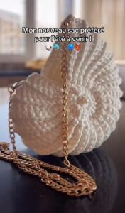 Sac crocheté à la main en coton recyclé, forme inspirée des coquillages, avec une chaîne en métal et une perle nacrée en ornement.