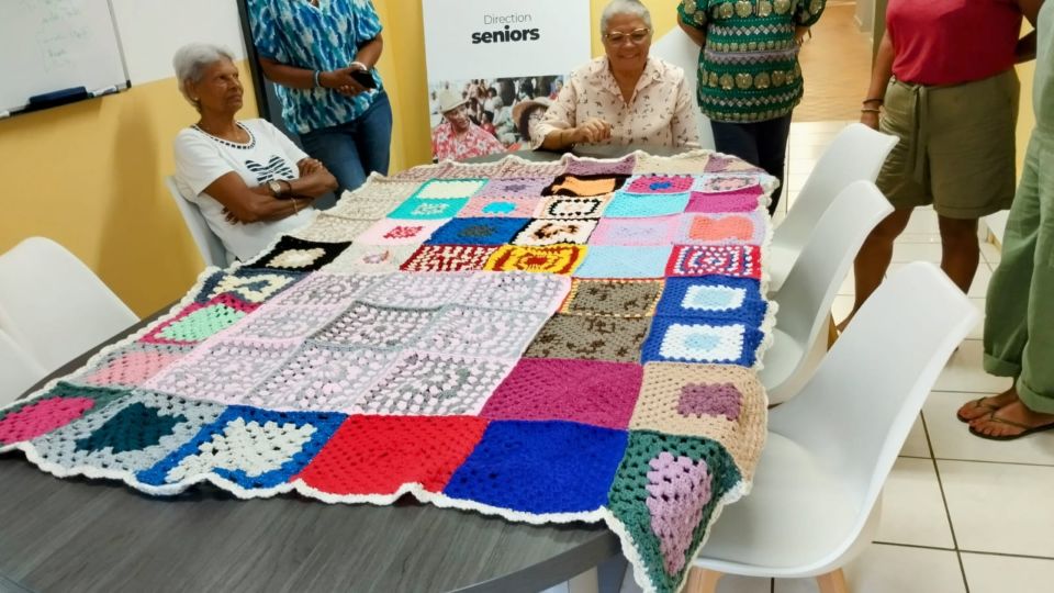 Photo d’un groupe de seniors souriantes en plein atelier tricot/crochet à la mairie annexe de La Providence. Des pelotes colorées, des ouvrages en cours, et une ambiance conviviale. Un plaid composé de carrés granny est visible en fond ou entre les mains des participantes.